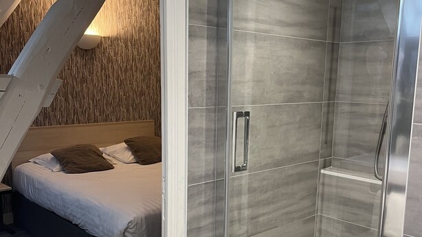 A modern bathroom with a glass shower enclosure, a wooden ceiling, and a bed with white bedding and brown pillows. - Hôtel De Troyes   (Troyes)