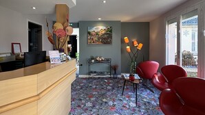 A reception area with a wooden counter, a floral carpet, red chairs, and a painting on the wall. - Hôtel De Troyes   (Troyes)