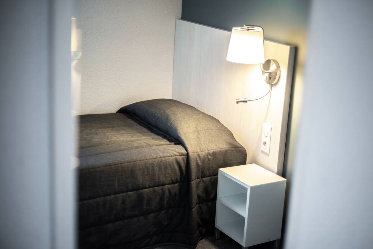 En pænt redt seng med et gråt tæppe, et hvidt natbord med en lampe og en stikkontakt.