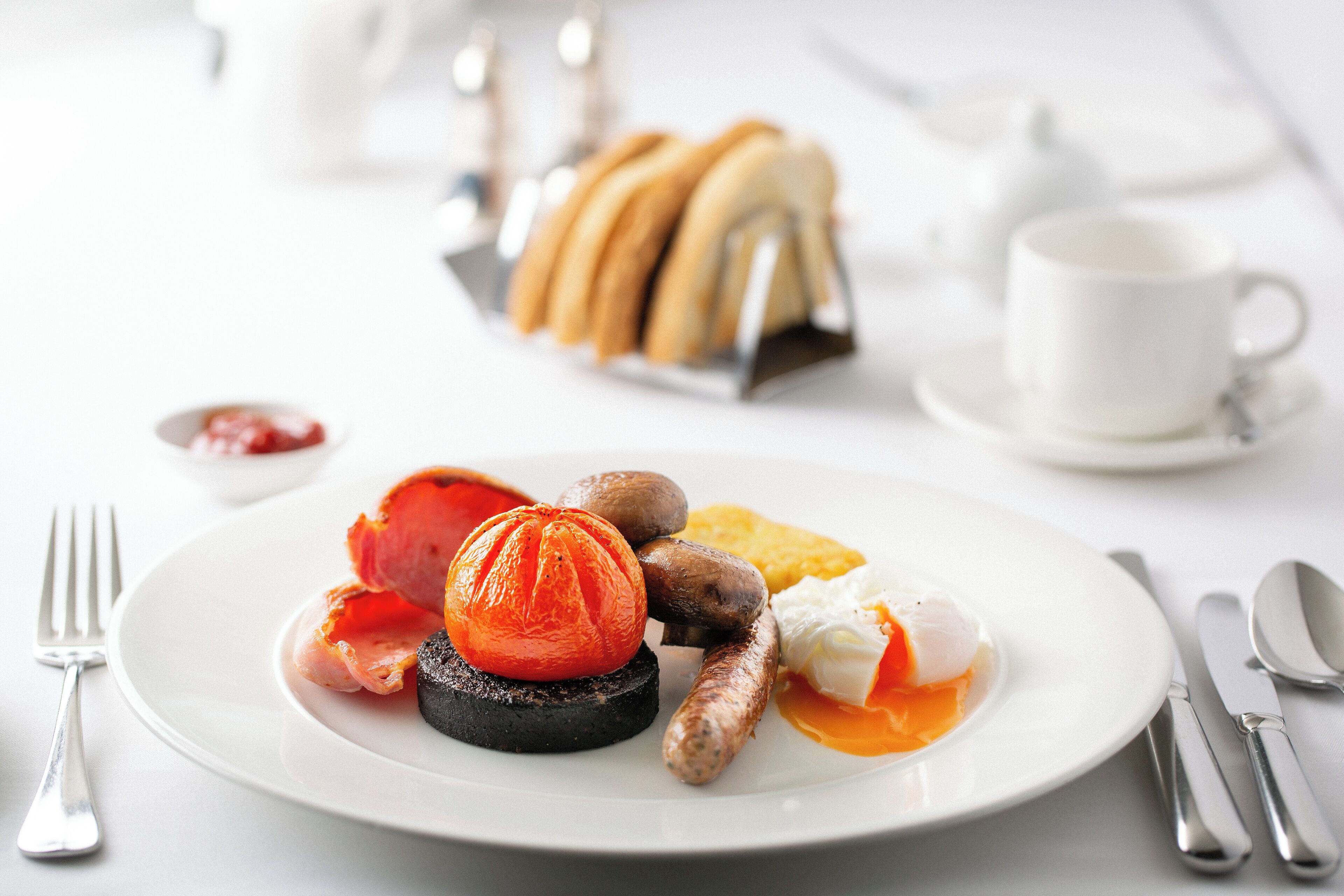 A breakfast plate with eggs, sausage, mushrooms, and a tomato on a black base, accompanied by a cup of coffee and a plate of croissants.