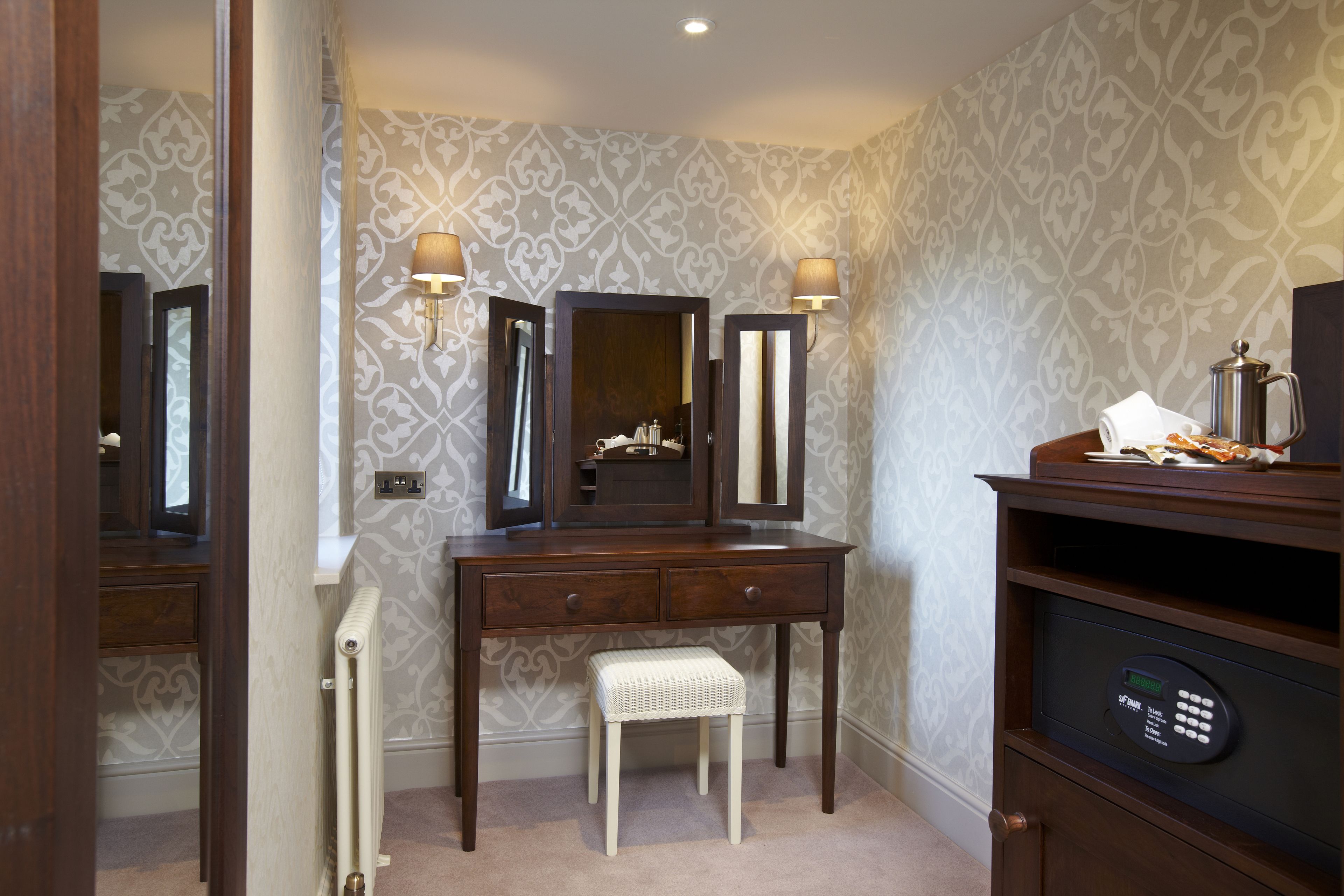 A bathroom with a vanity, two wall-mounted mirrors, a radiator, a bench, and a shelf with a coffee pot.