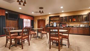 A hotel dining area with wooden tables and chairs, a kitchen area with cabinets and appliances, and a large window with curtains. - Best Western Plus Jacksonville West (Jacksonville)