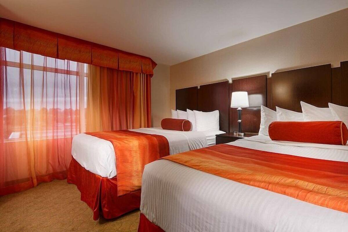 A hotel room with two beds, a nightstand, a lamp, and a large window with curtains.