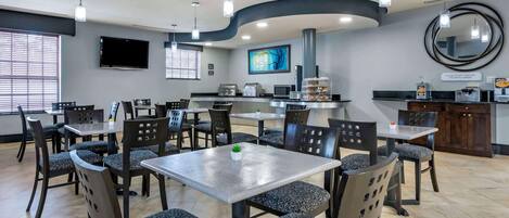 A modern restaurant with a central dining area, a kitchen, and a bar. There are multiple tables and chairs, a flat-screen TV, and a large mirror on the wall.