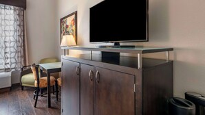 A hotel room with a flat-screen TV mounted on a glass shelf above a dark wooden cabinet with two doors. There is a table with two chairs and a lamp on the table. A painting is visible on the wall. - Best Western Plus Bradenton Gateway Hotel (Bradenton)