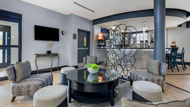 A modern lounge area with a round black coffee table, patterned armchairs, a flat-screen TV, and a bar area with a coffee machine. - Best Western Plus Bradenton Gateway Hotel (Bradenton)