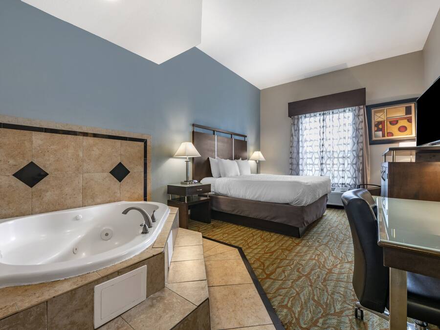 A hotel room with a large bathtub, a bed with two pillows, a desk with a flat-screen TV, and a window with patterned curtains.