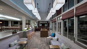 Un hall d’entrée spacieux avec un espace salon central, une cuisine à l’arrière-plan et de grandes fenêtres sur le côté droit.