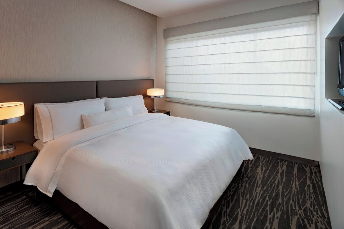 A hotel room with a large bed, two bedside tables with lamps, a window with blinds, and a carpeted floor.