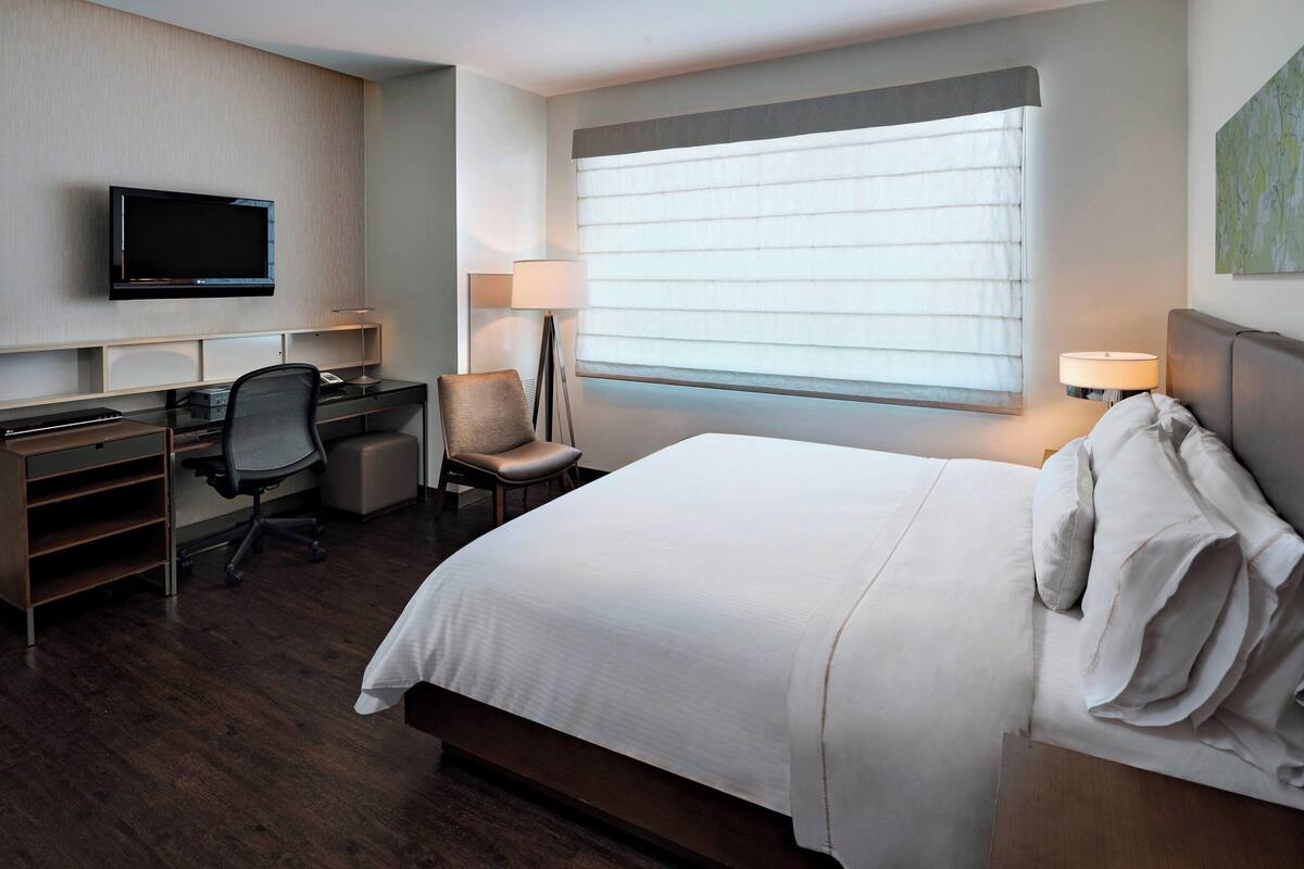 A hotel room with a bed, desk, chair, TV, and window with blinds.