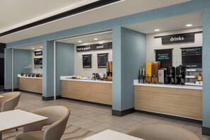 A modern cafe with a counter displaying hot foods, fruit & yogurt, and cereal. There are seating areas with tables and chairs. - Holiday Inn Columbus-Hilliard by IHG (Columbus)