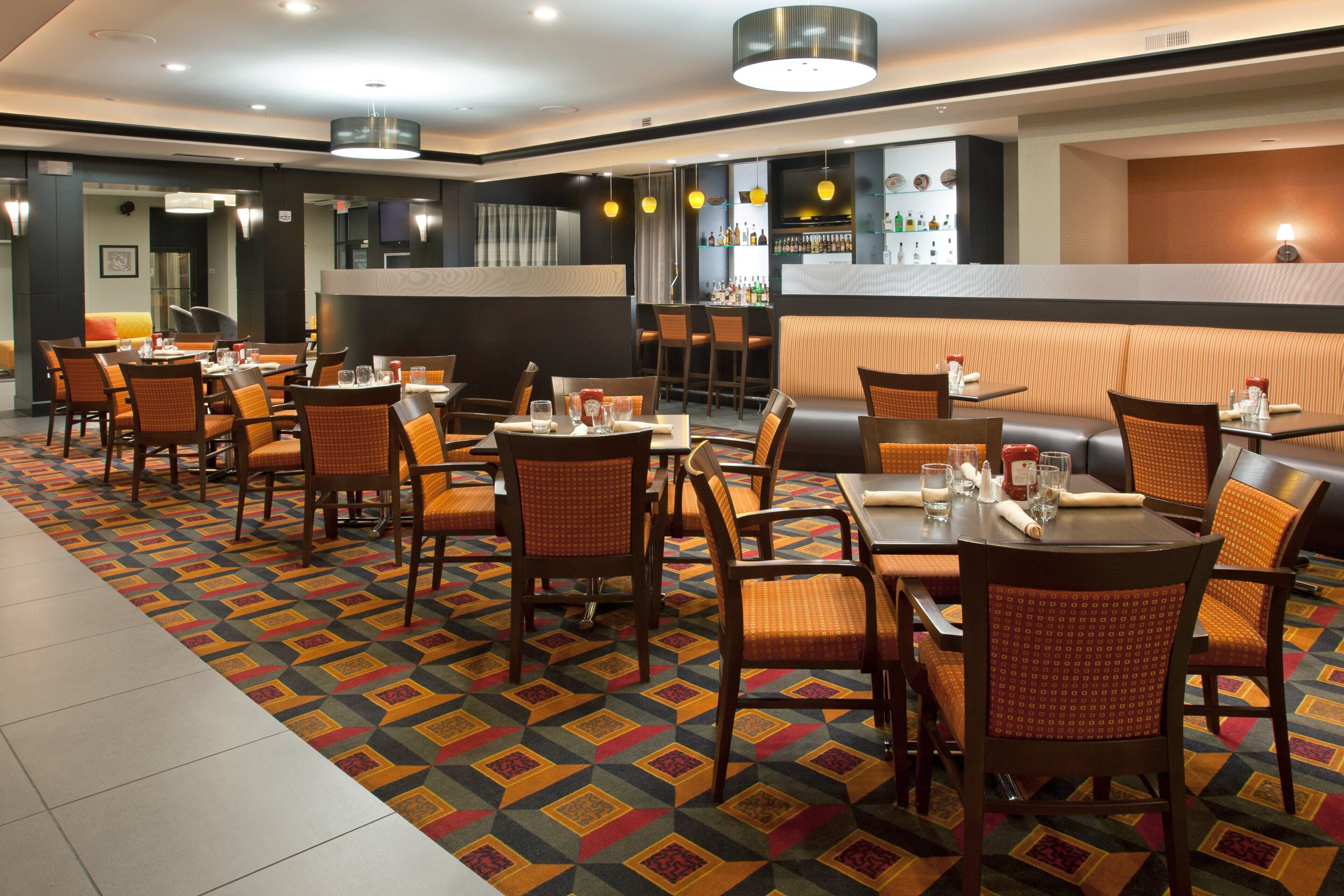A hotel dining area with multiple tables and chairs, a patterned carpet, and a bar area in the background.