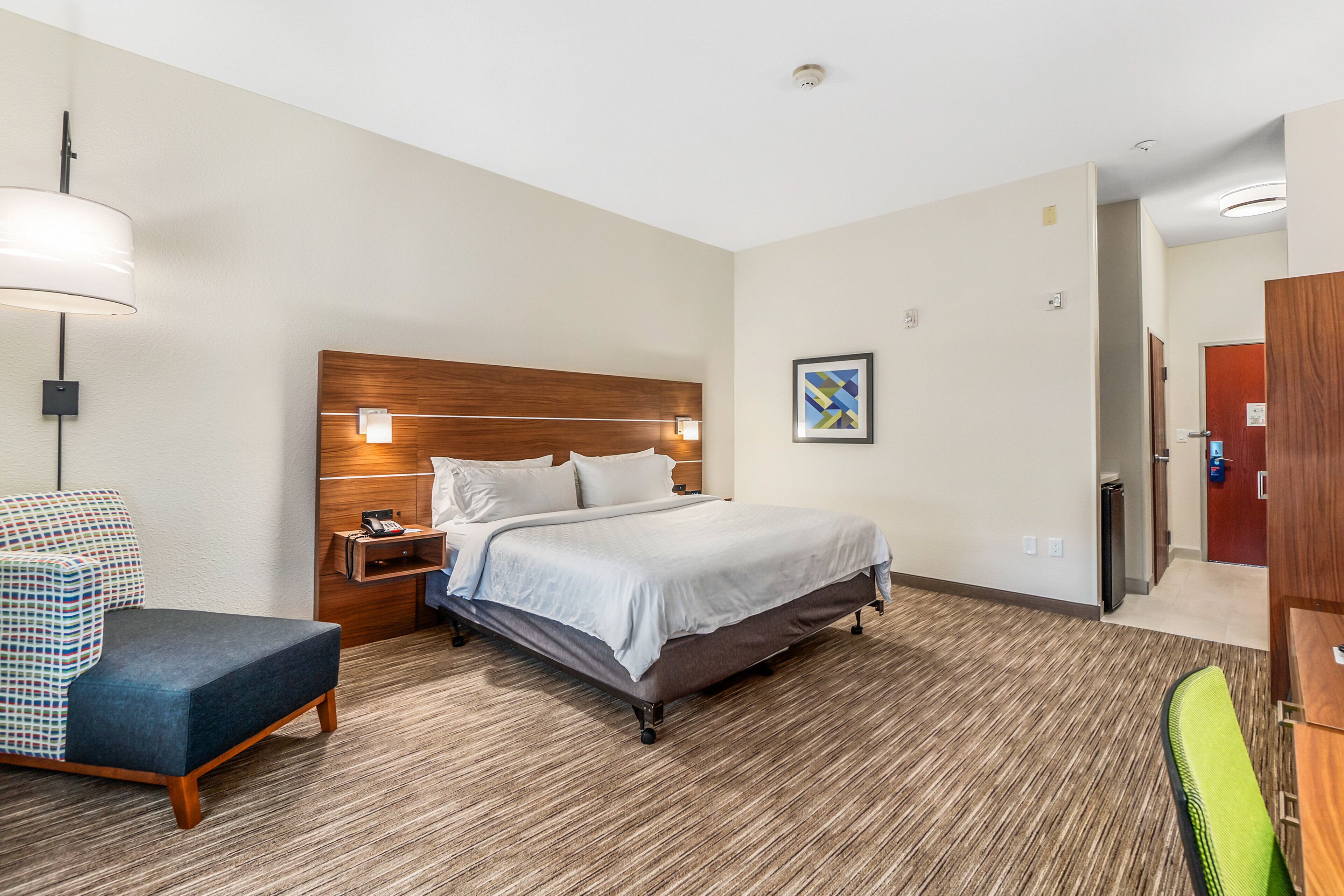 A hotel room with a bed, a desk, a chair, a lamp, and a painting on the wall.