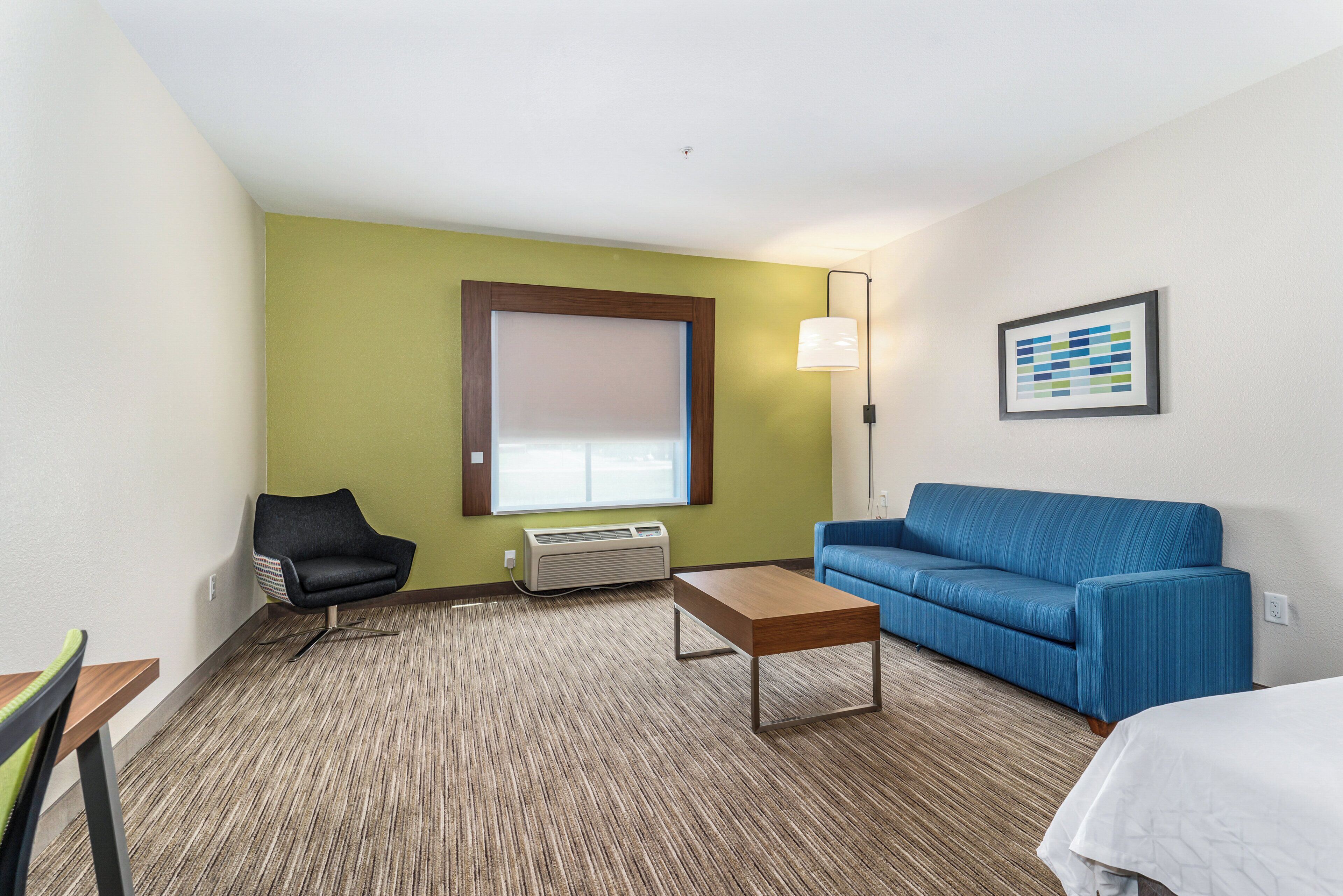 Ein Hotelzimmer mit einem blauen Sofa, einem Holztisch, einem schwarzen Sessel, einem Fenster mit Jalousien, einer Stehlampe und einem gerahmten Bild an der Wand.