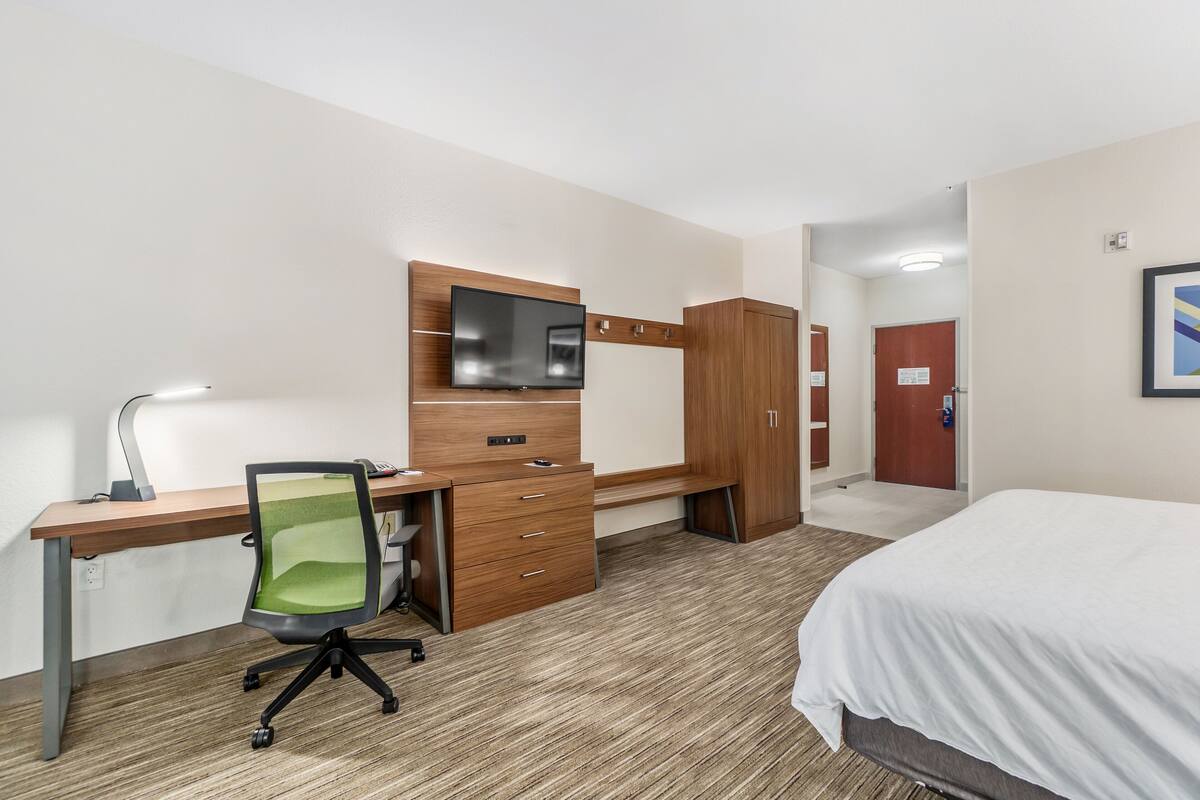 A hotel room with a wooden desk, a green office chair, a bed, and a television mounted on the wall.