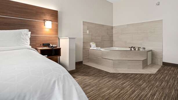 A modern hotel room with a large bed, a wooden headboard, a built-in desk with a telephone, and a corner bathtub with a towel and toiletries. - Holiday Inn Express Suites Van Buren-Ft Smith Area by IHG (Van Buren)