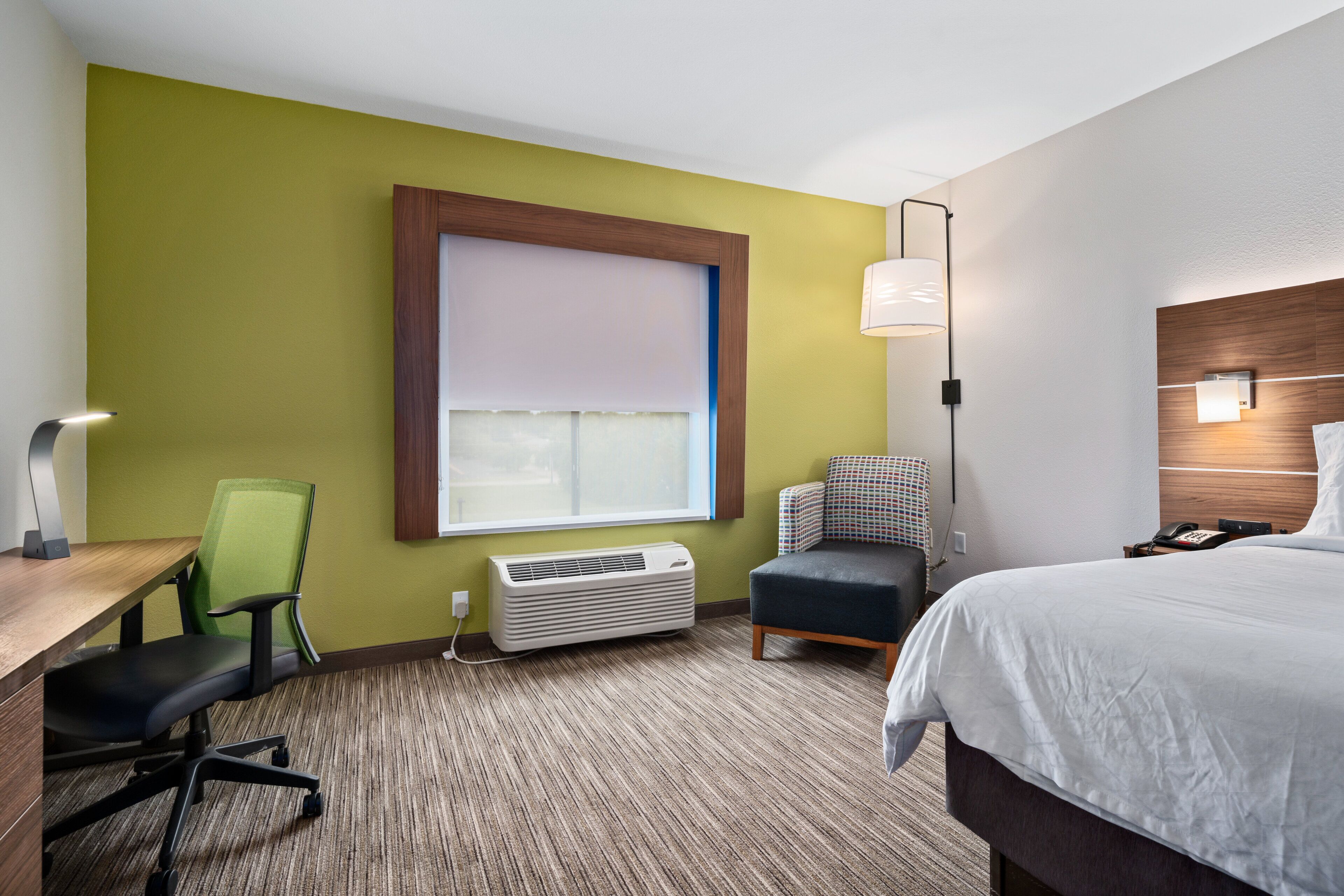 Habitación de hotel con cama, escritorio con silla, ventana con persianas, lámpara de pared y aire acondicionado instalado en la pared.