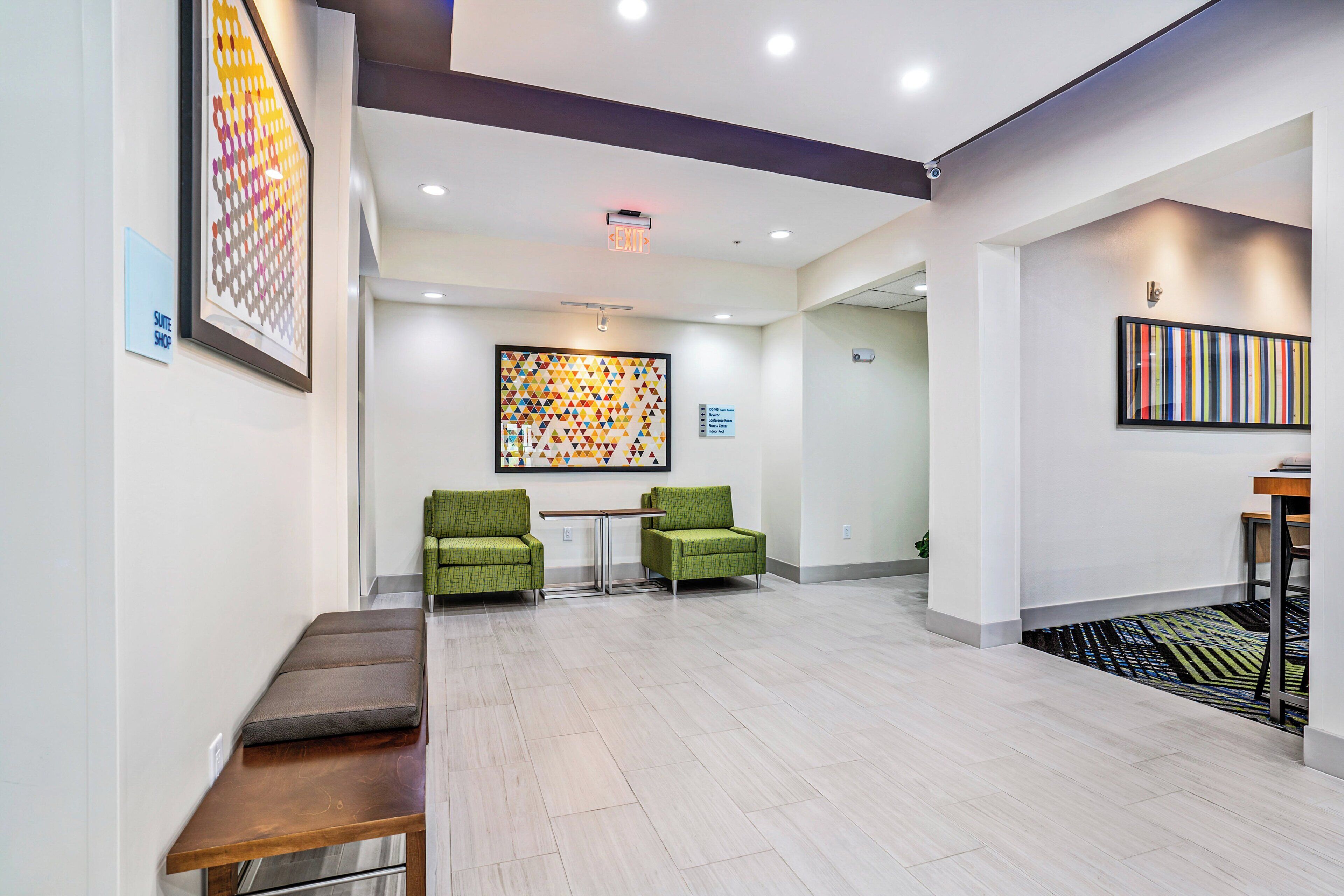 A hotel lobby with a reception desk, two green armchairs, a bench, and a colorful abstract painting on the wall.