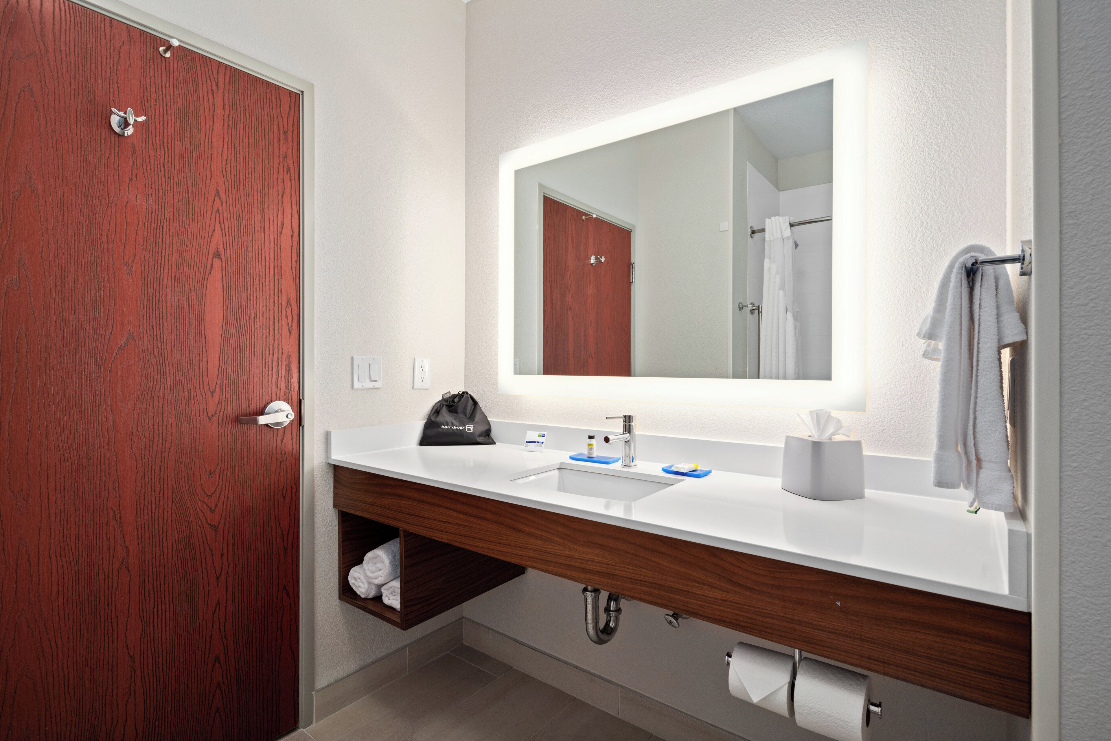 Un baño moderno con puerta de madera, un lavabo blanco, un espejo con luz y un perchero para toallas.
