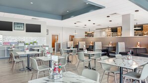 Un restaurant moderne avec des tables surmontées de marbre, des chaises grises et un coin bar agrémenté d’un mur en pierre.