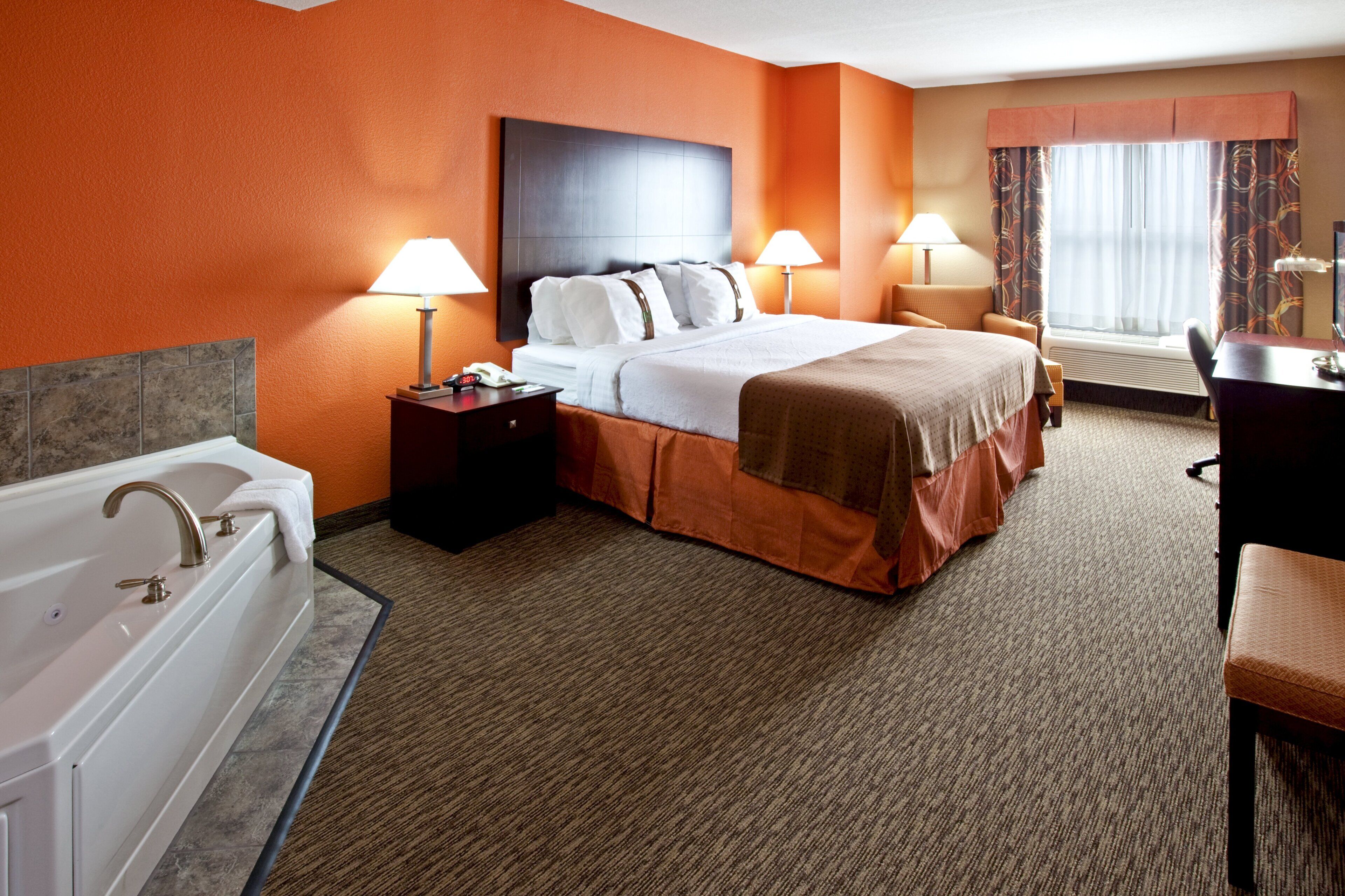 A hotel room with a large bed, two bedside tables with lamps, a desk, a chair, a bathroom with a bathtub, and a window with curtains.