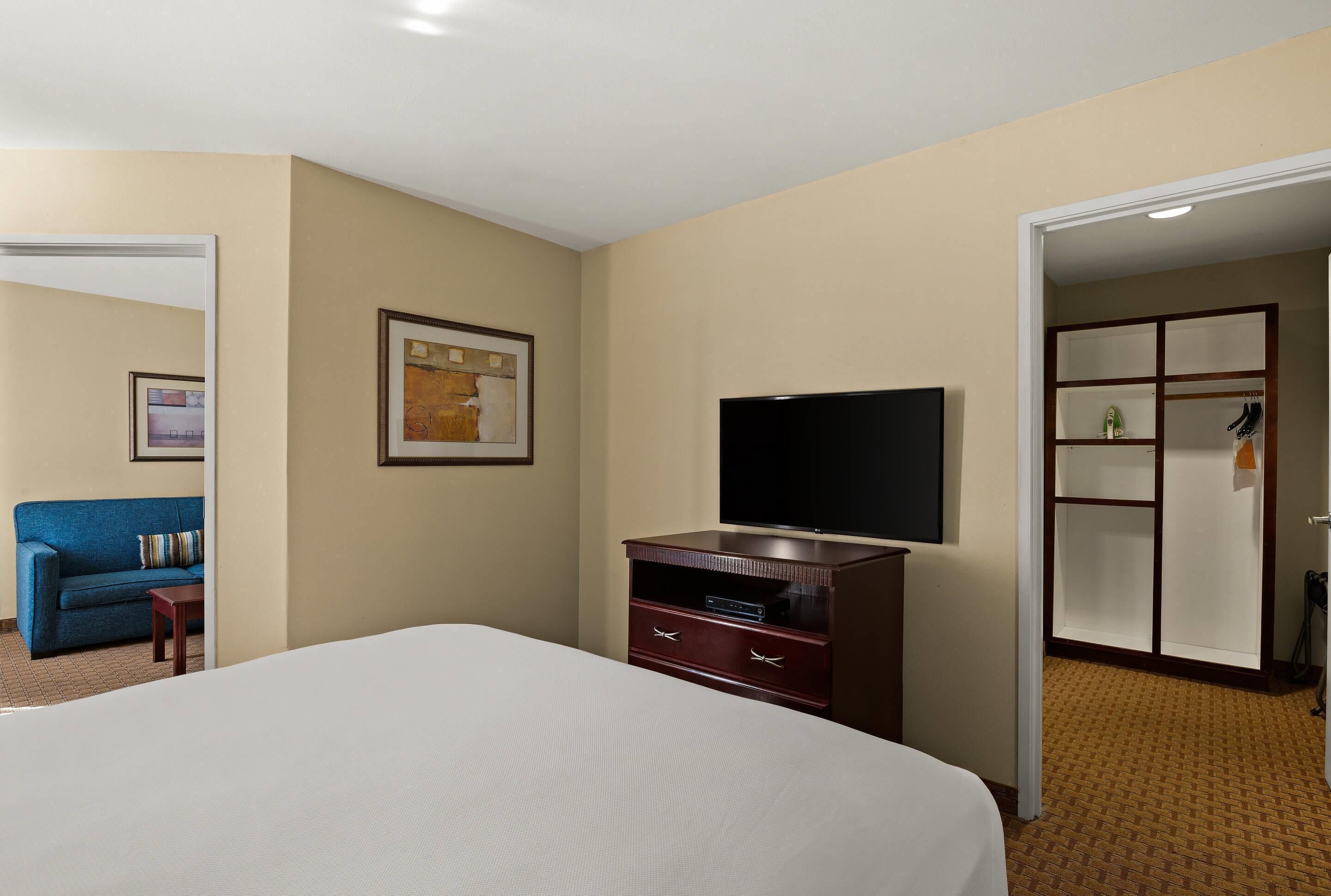 Una habitación de hotel con una cama, televisión, cómoda, un cuadro enmarcado y una puerta que da a otra habitación.
