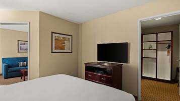Una habitación de hotel con una cama, televisión, cómoda, un cuadro enmarcado y una puerta que da a otra habitación.