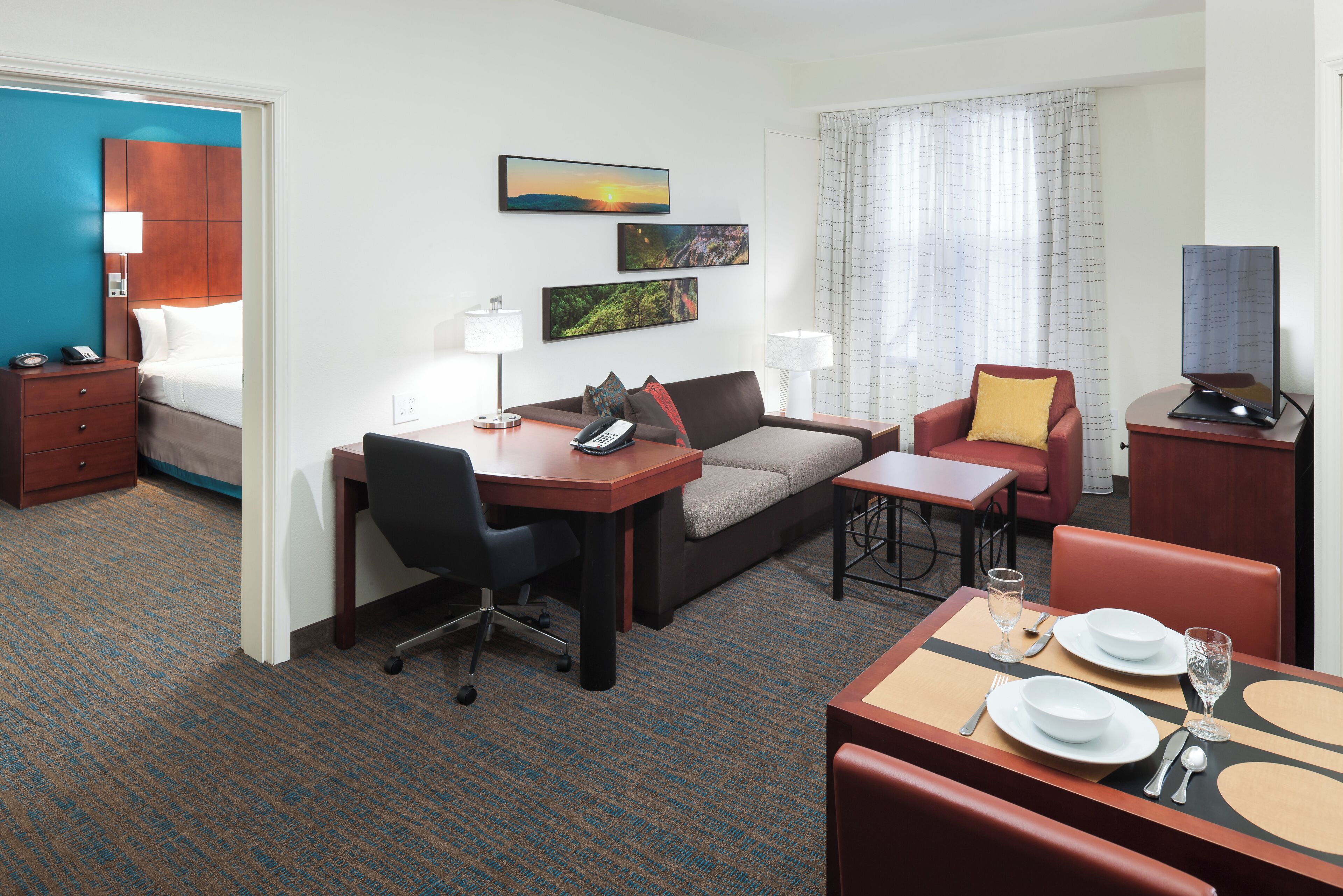 A hotel room with a bed, a desk, a sofa, a dining table set, and artwork on the walls.