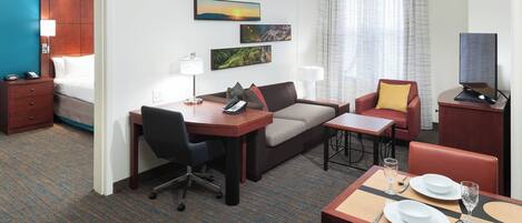 Ein Hotelzimmer mit Bett, Schreibtisch, Sofa, Esstisch für zwei Personen und Fernseher.
