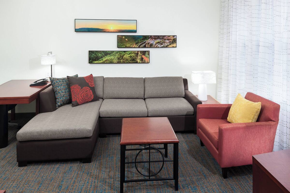 Una sala de estar moderna con un sofá seccional gris, un sillón rojo y una pequeña mesa de madera. Hay tres cuadros enmarcados en la pared, una lámpara en una mesita auxiliar y una alfombra estampada.