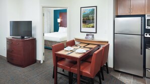 A hotel room with a dining table set for two, a refrigerator, a television, and a bed with a view.