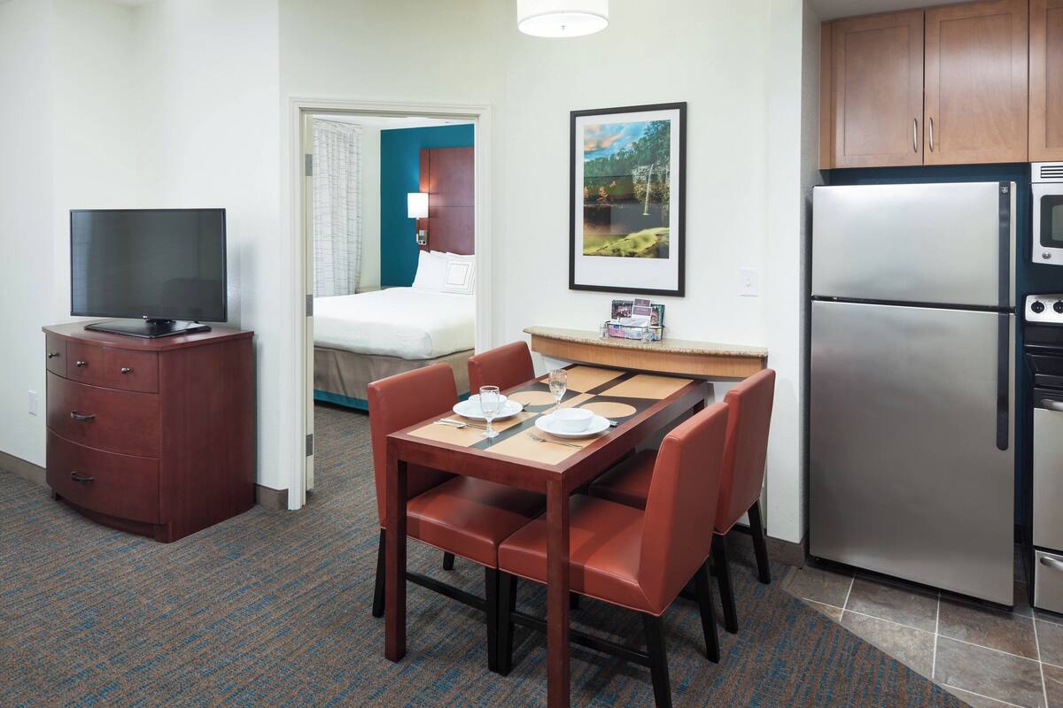 Habitación de hotel con mesa de comedor para dos personas, refrigerador, televisión y cama con vistas.