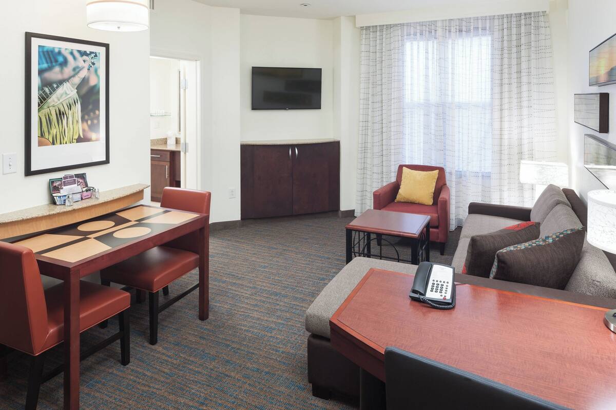 Habitación de hotel con mesa de comedor, dos sillas, un sofá, un televisor y una ventana con cortinas.