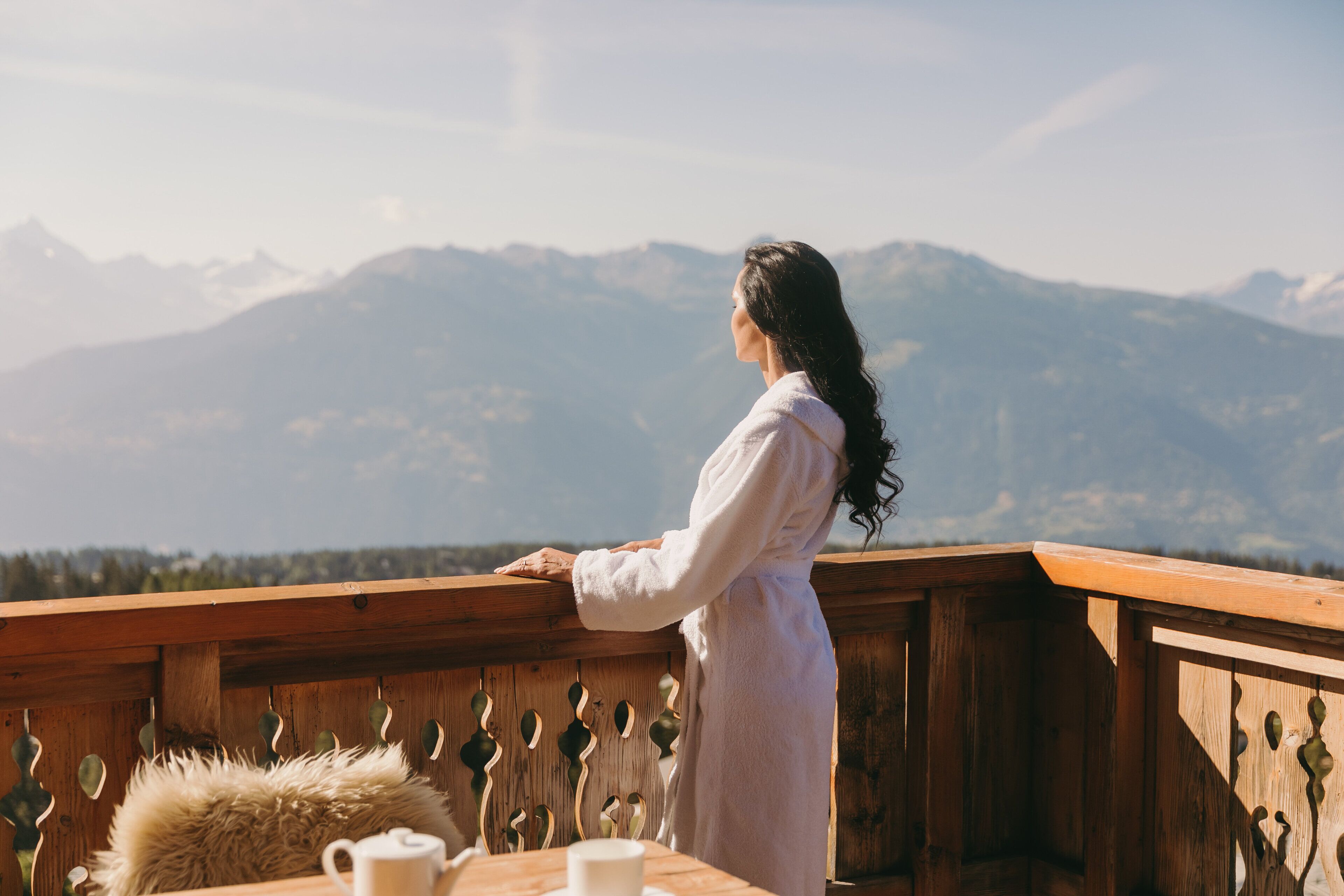 Uma pessoa vestida com um roupão branco está em um varanda de madeira com vista para uma paisagem montanhosa.