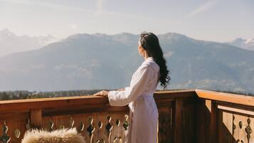 Uma pessoa vestida com um roupão branco está em um varanda de madeira com vista para uma paisagem montanhosa.