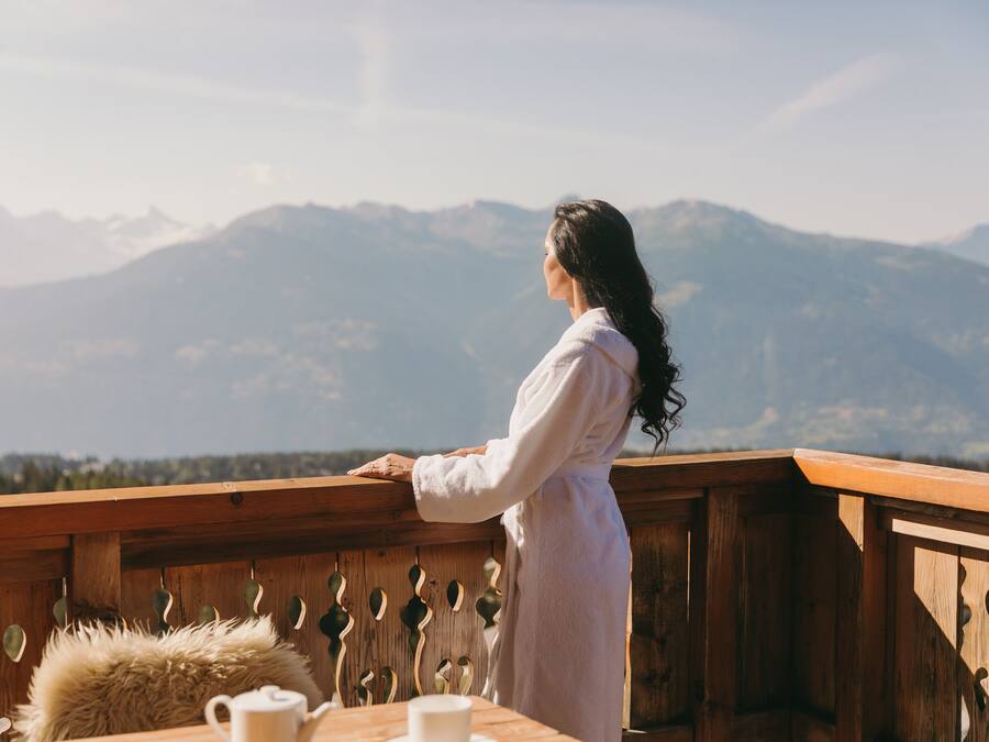 Uma pessoa vestida com um roupão branco está em um varanda de madeira com vista para uma paisagem montanhosa.