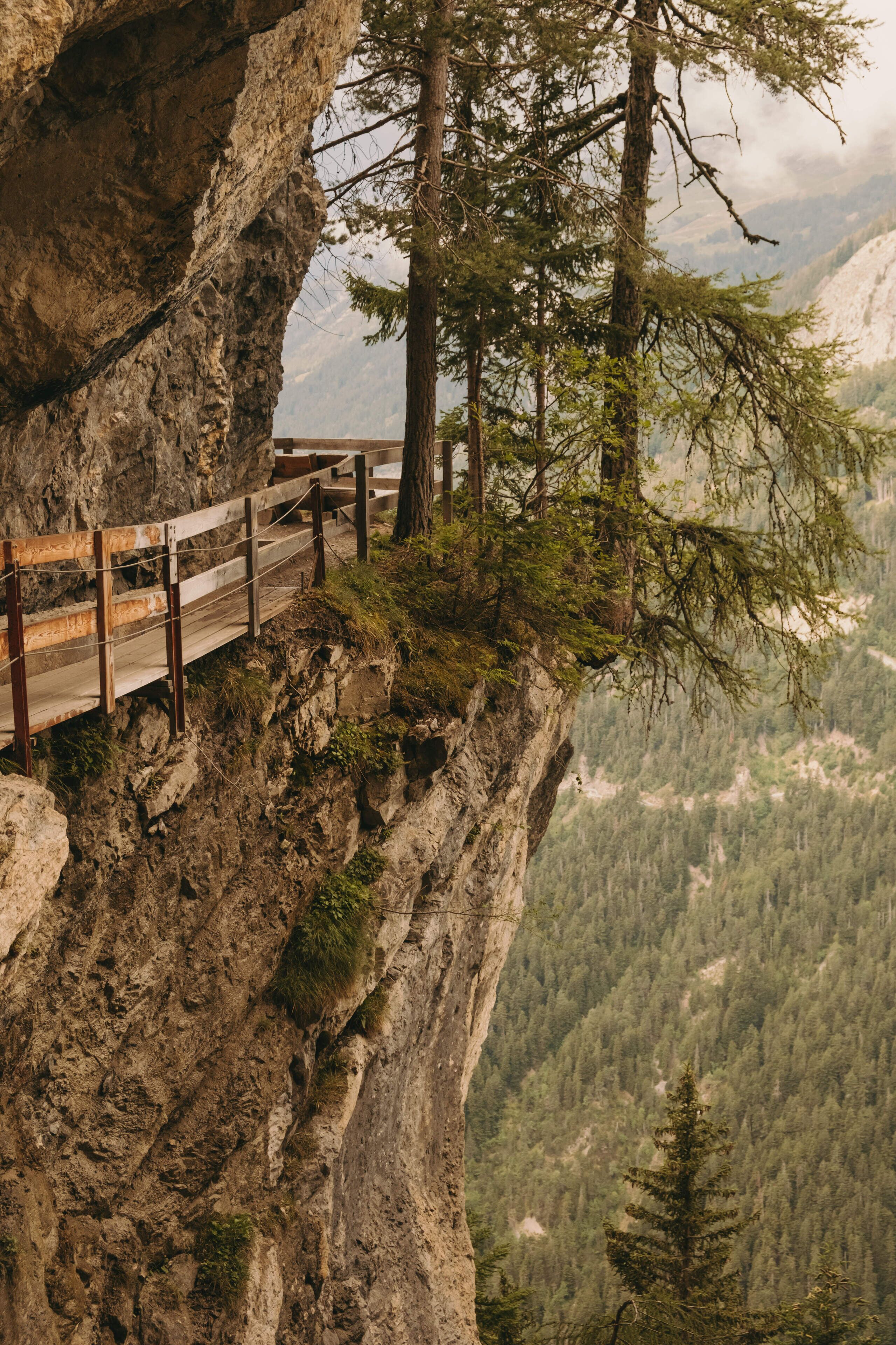 Un pont en bois, doté de garde-corps, est construit sur une falaise rocheuse, d’où l’on peut admirer une vallée boisée.
