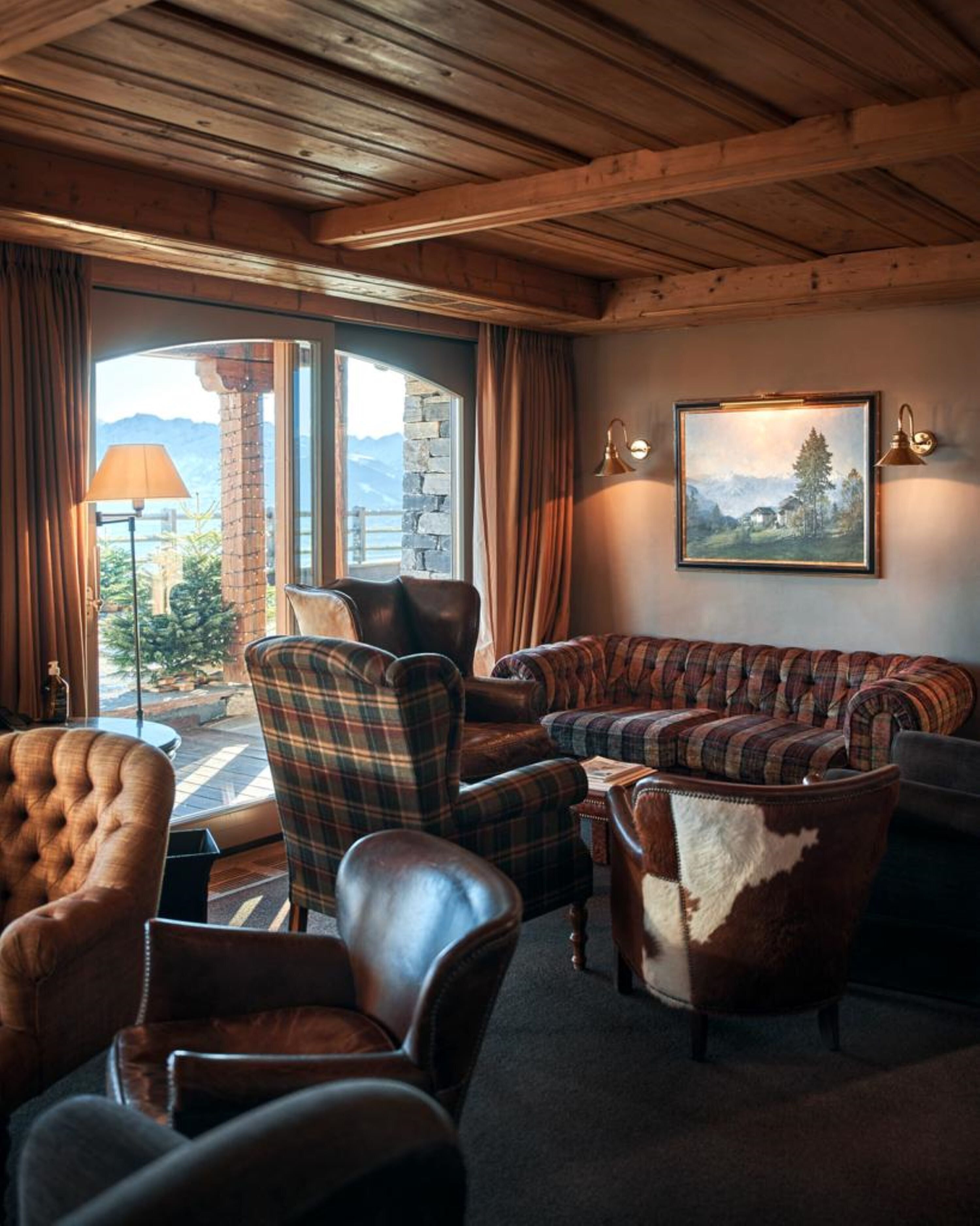 Un salon chaleureux avec un canapé, des fauteuils et une cheminée. Une grande fenêtre offre une vue sur les montagnes et un lac.