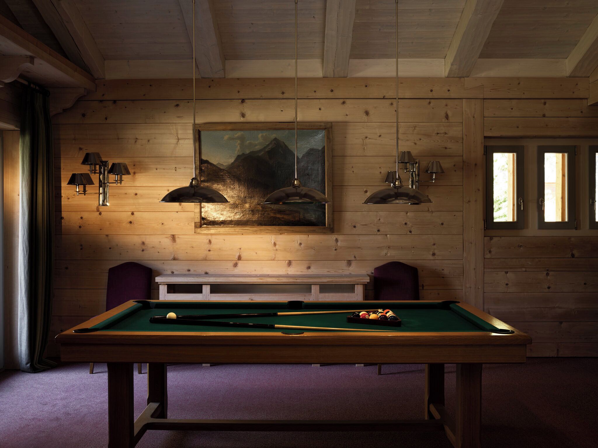 A pool table in a room with wooden walls, a painting above it, and two lamps on the wall.