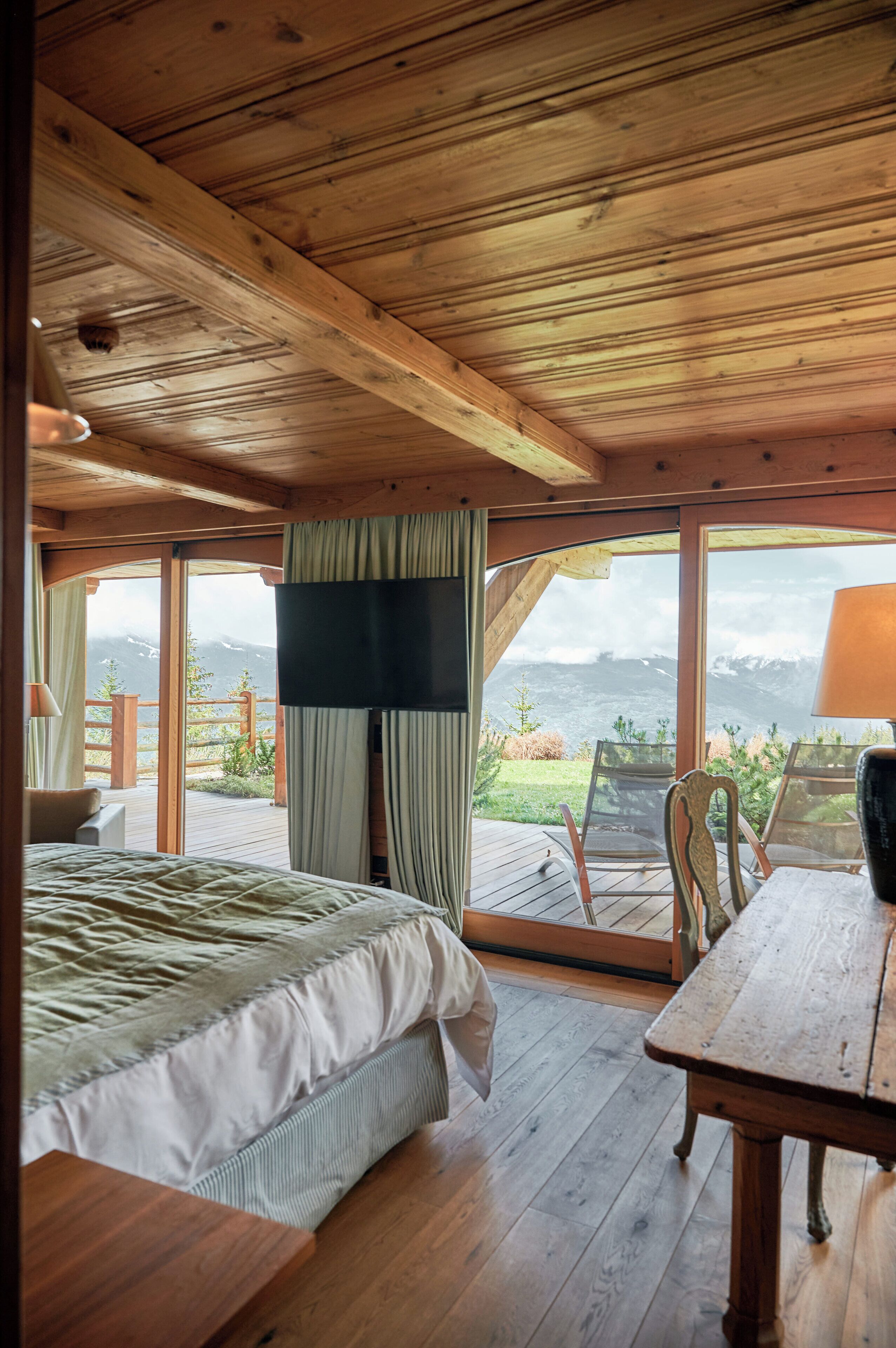 A wooden cabin room with a bed, a table, and a view of mountains through large windows.