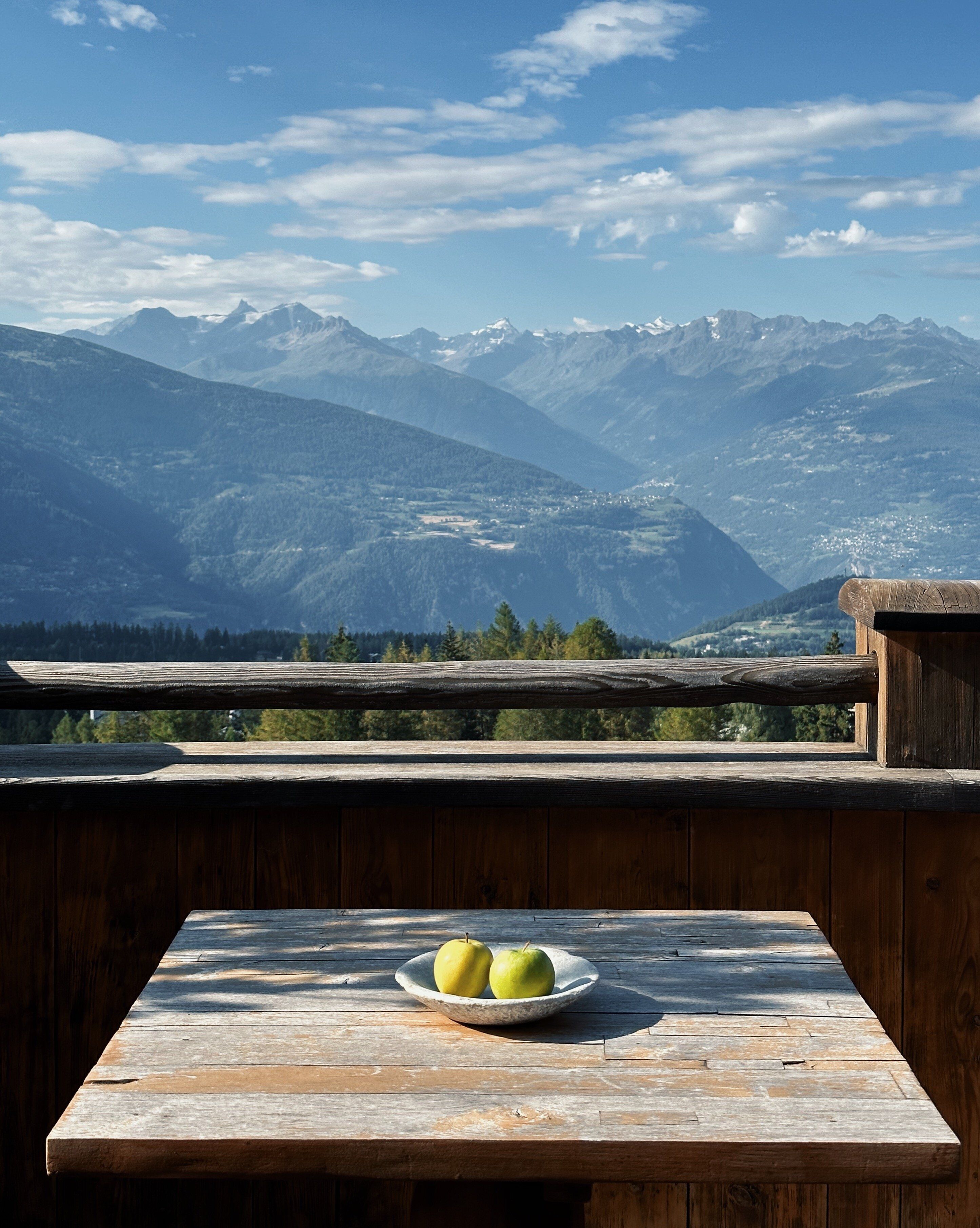 A wooden table with a bowl of apples, overlooking a mountainous landscape.
