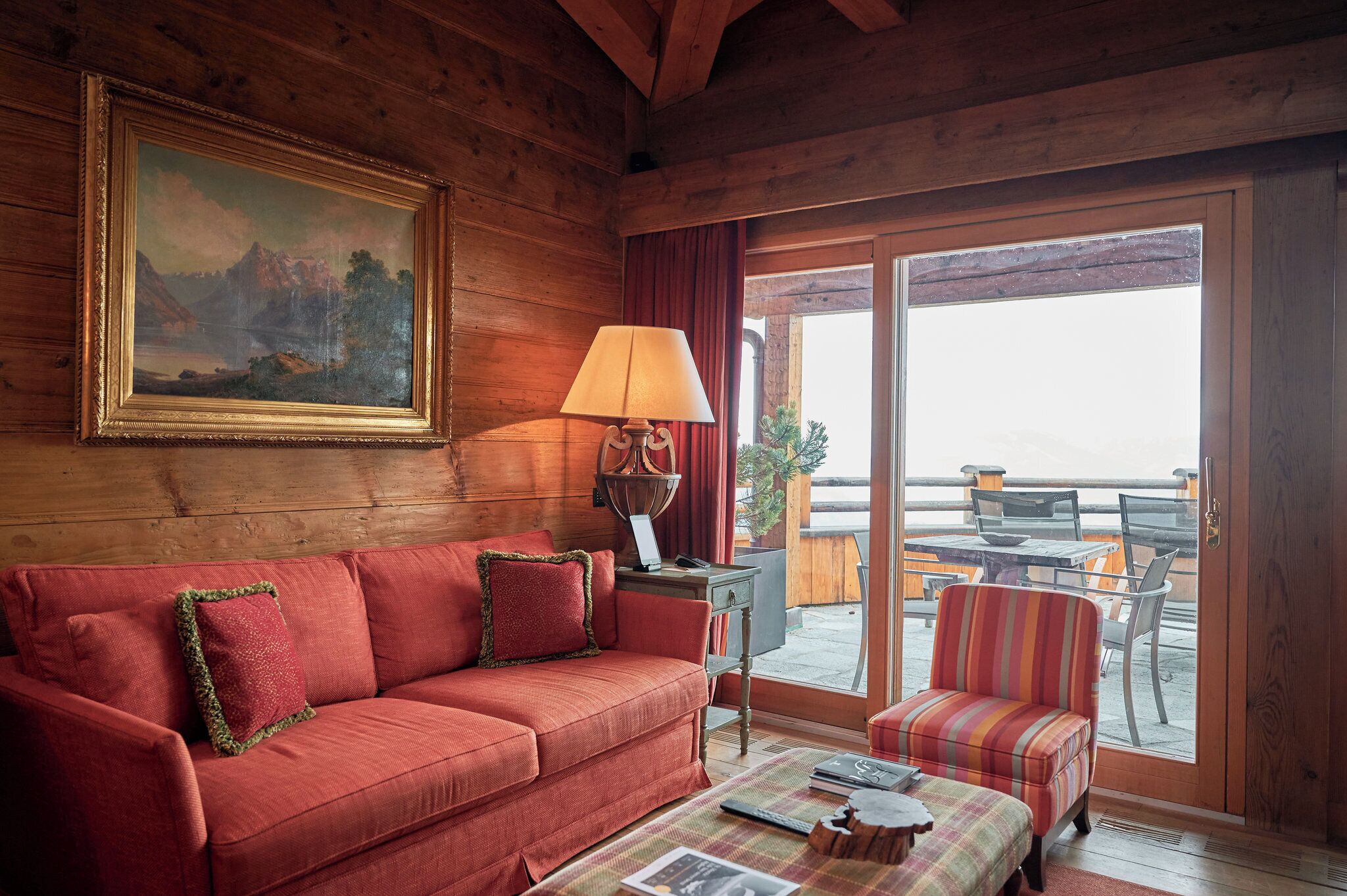 A wooden interior with a sofa, a lamp, a painting, and a view through a glass door to an outdoor seating area.