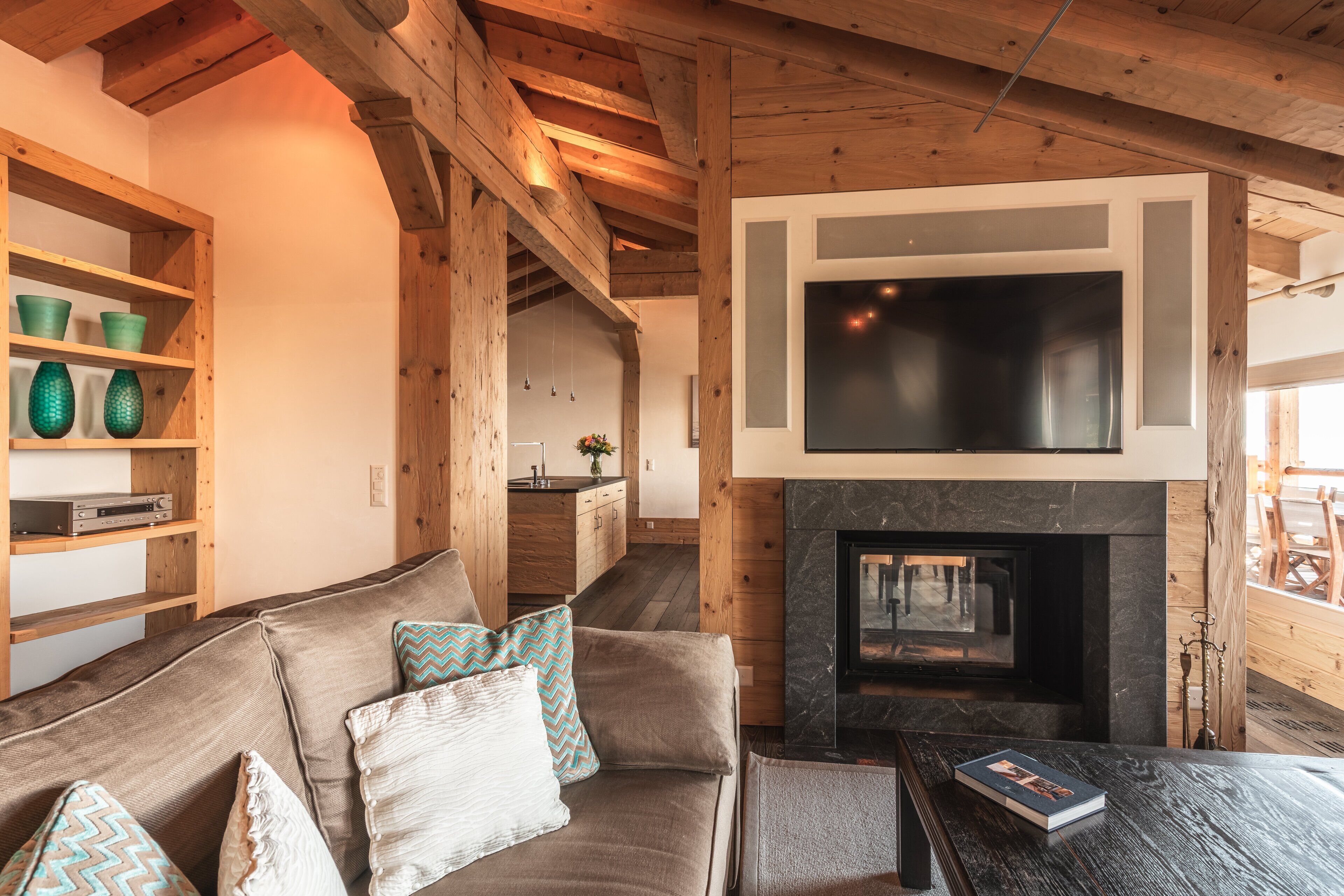 Ein gemütliches Wohnzimmer mit Kamin, Holzbalken, einer Couch mit gemusterten Kissen und einem flachbildfernseher, der über dem Kamin angebracht ist.