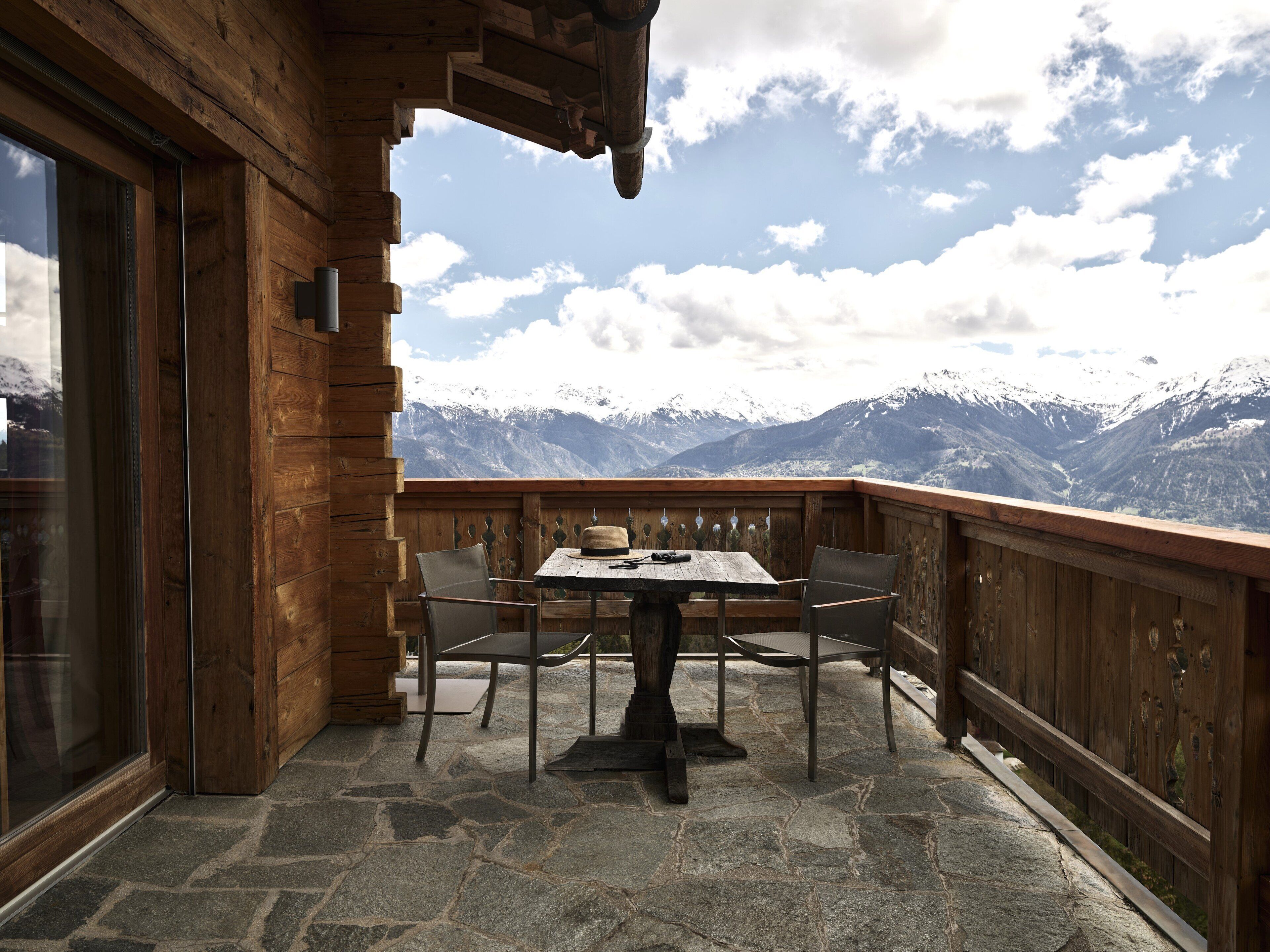 Una terraza de madera con suelo de piedra, una mesa y sillas, con vistas a un paisaje montañoso.