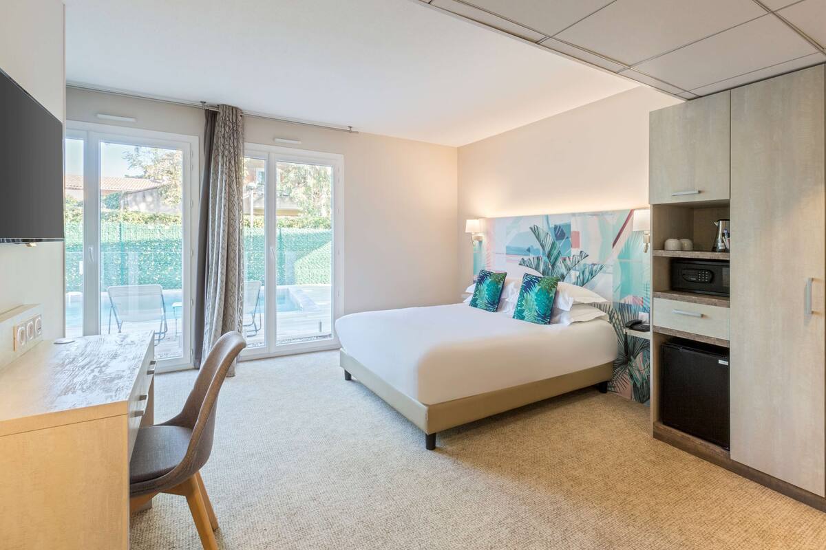 A modern hotel room with a large bed, a desk with a chair, a flat-screen TV, a small refrigerator, and a sliding glass door to the outside.
