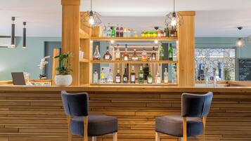 Zona de bar con estantes de madera llenos de botellas, dos taburetes y lámparas colgantes.