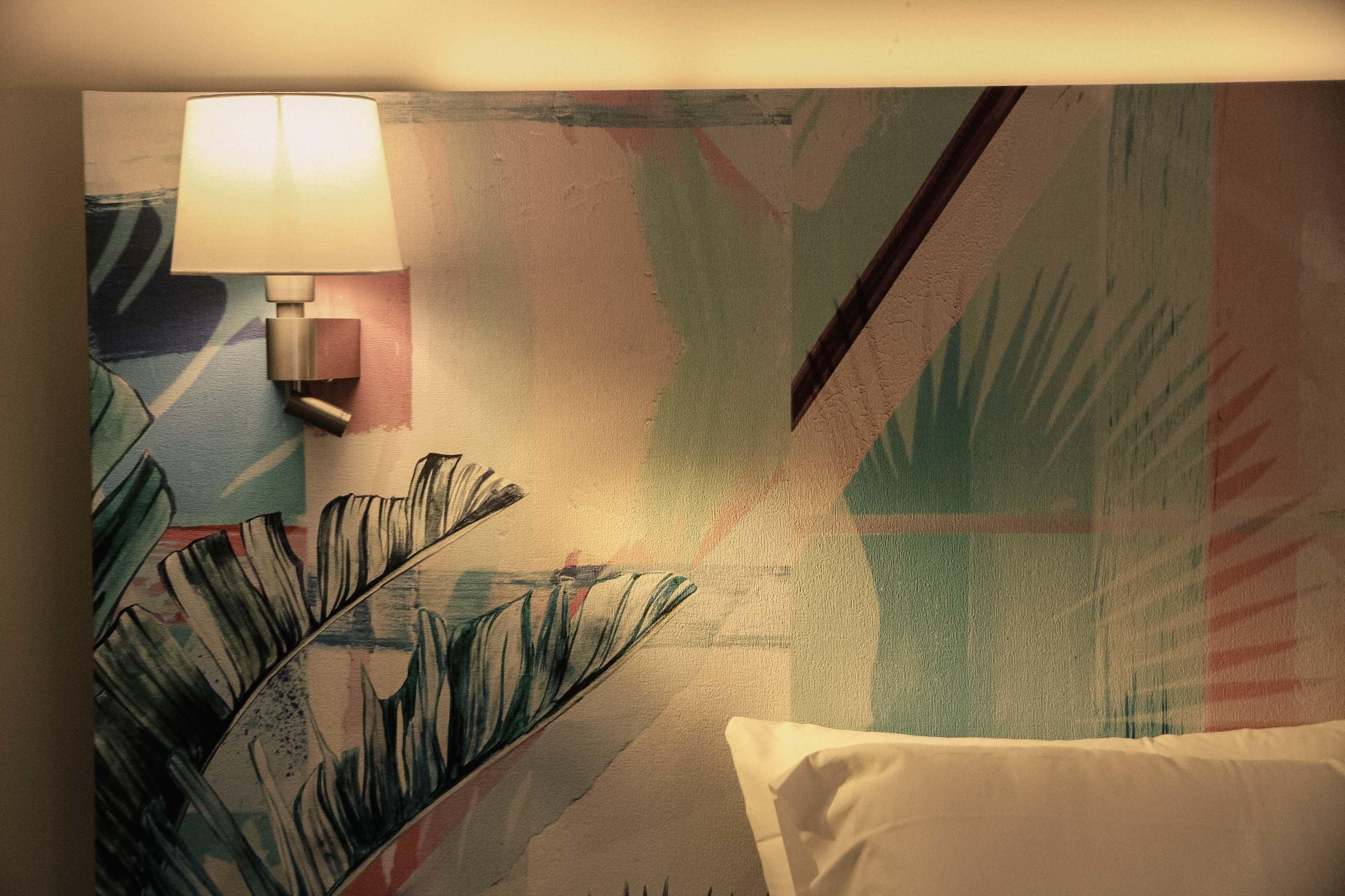 Una habitación de hotel con una cama, una lámpara fijada a la pared y un cuadro de una hoja tropical grande.
