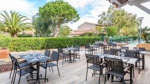 Un patio al aire libre con mesas y sillas, rodeado de vegetación y un edificio al fondo.