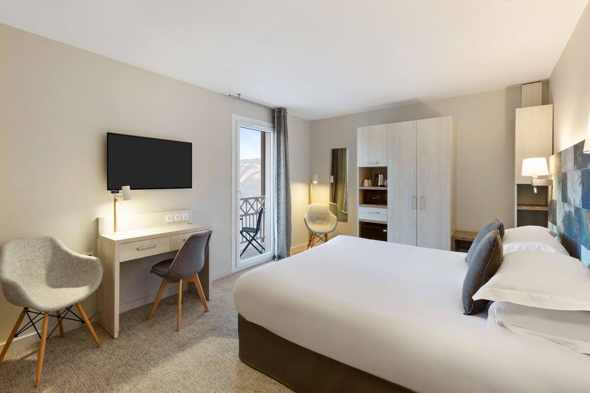 A hotel room with a bed, desk, chair, TV, wardrobe, and a balcony with a view.