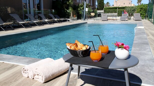 Escena junto a la piscina con sillones de descanso, una mesa con cruasanes, zumo de naranja y un jarrón con flores.